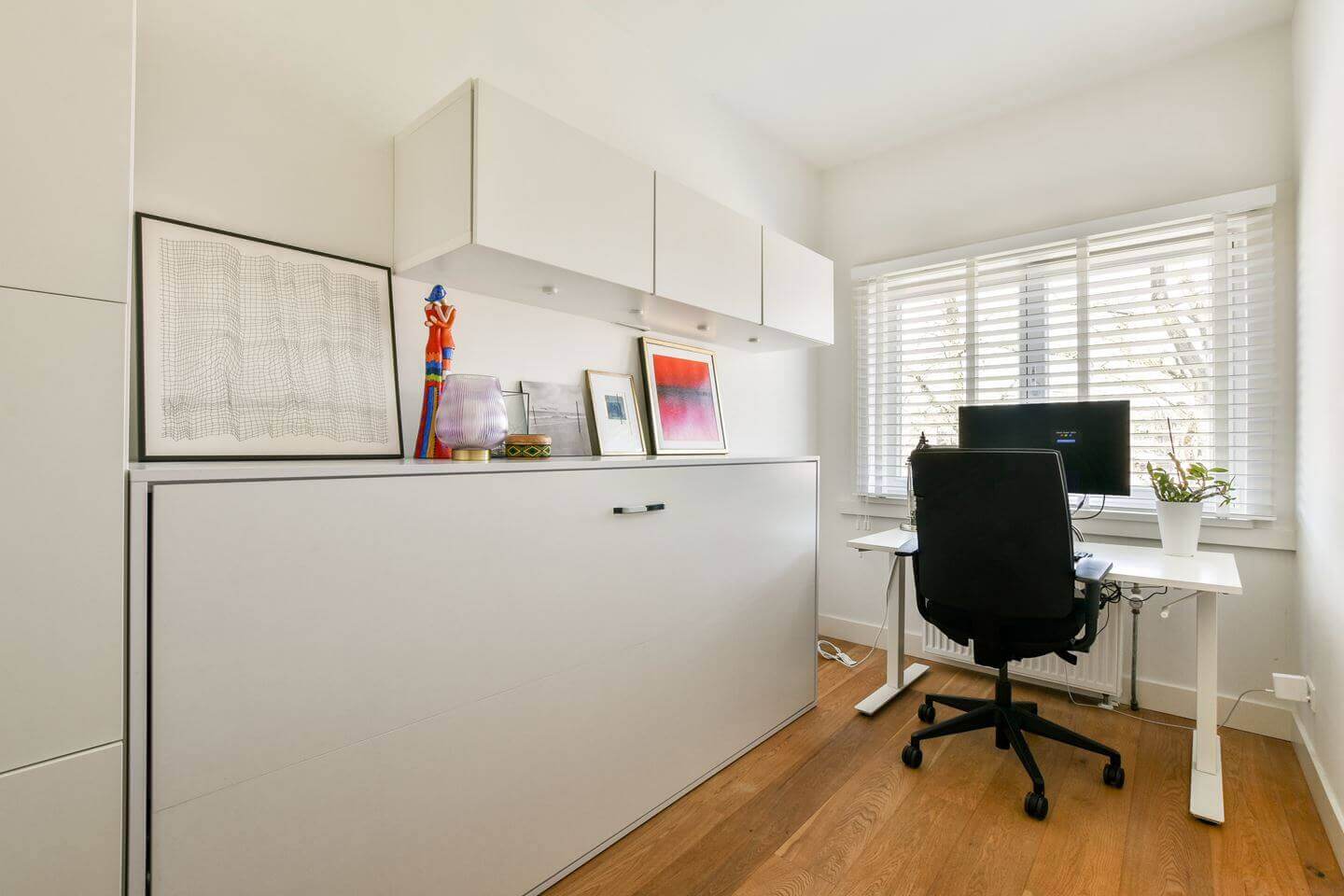 Modern thuiskantoor met een wit opklapbed, bureau en computer, ideaal voor ruimtebesparing en comfort in een kleine ruimte.